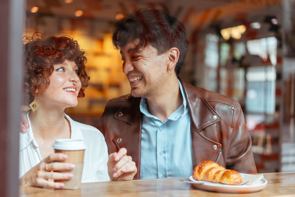 A young couple sitting close together at a coffee shop table, smiling during conversation